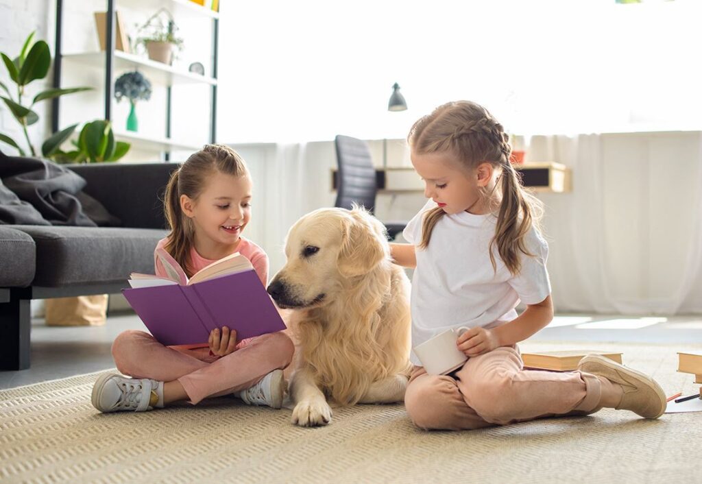 two little girls sitting on floor reading to Labrador retriever dog